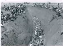Black-and-white photo showing large numbers of Kenyan natives digging a deep canal using hoes and shovels during the Mau Mau emergency in colonial Kenya. Workers crowd along the trench.