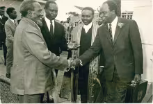 Black-and-white photo showing President Jomo Kenyatta shaking hand with Finance minister Mwai Kibaki wearing a dark suit, white tie and patterned tie, and holding the budget briefcase with his left hand. At the background, between the two, are Vice President Daniel arap Moi and the first African Clerck of the National Assembly Leonard James Ngugi in his official parliamentary attire.