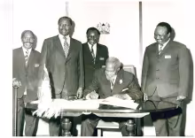 Black-and-white photo showing President Jomo Kenyatta signing an official document as his Vice Daniel Moi and other State officials stand round him all smiles. Kenyatta wears a dark suit, striped tie and flower lapel pin, while the officials are dressed in formal suits some with conference name badges. Kenyatta's ceremonial fly whisk rests on the table infront of him.